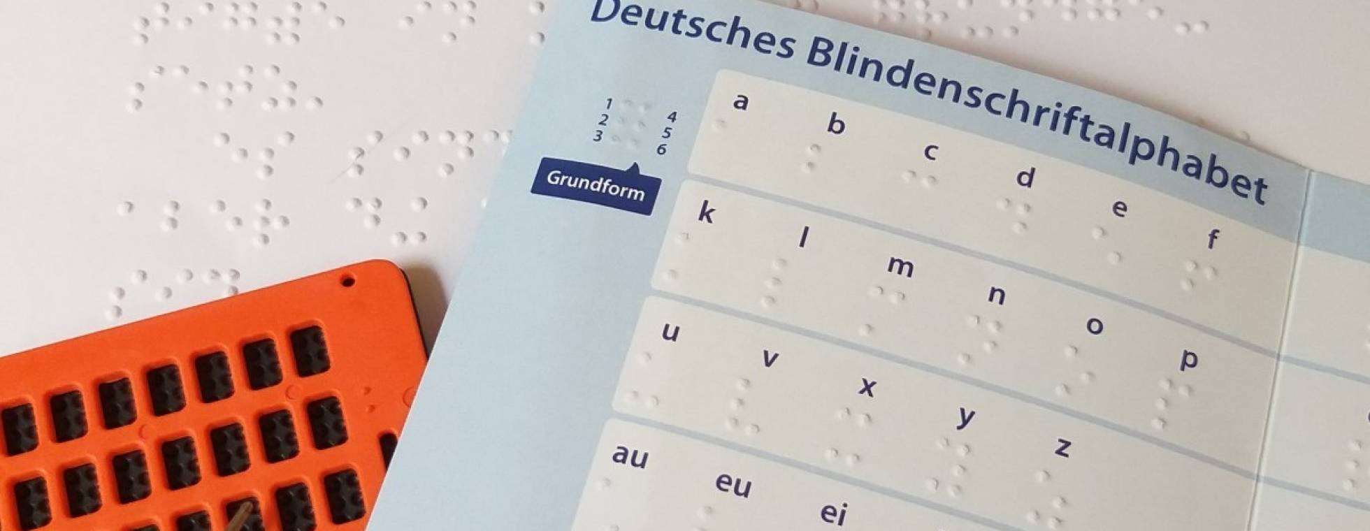 Vor einer Seite in Braille liegt ein aufgeschlagenes Braille-Alphabet und eine Punktschrift-Tafel mit Griffel