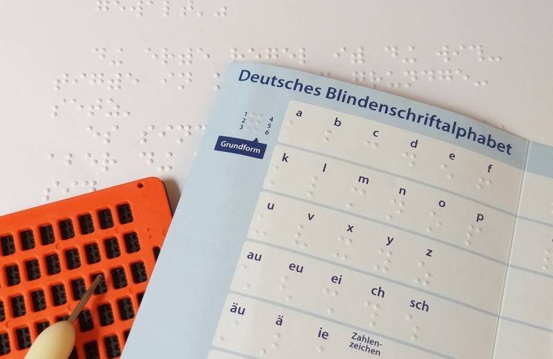 Vor einer Seite in Braille liegt ein aufgeschlagenes Braille-Alphabet und eine Punktschrift-Tafel mit Griffel