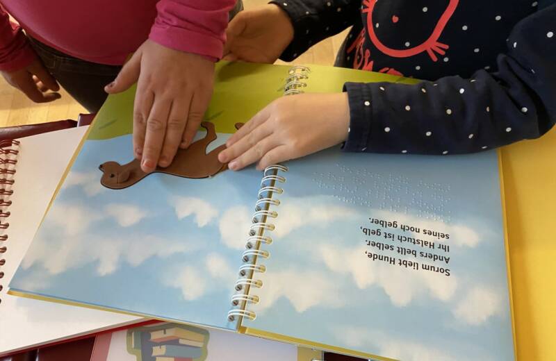Über weiteren Büchern ist ein Kinderbuch aufgeschlagen. Zwei Kinder stehen vor dem Buch, ihre Hände betasten eine taktile Abbildung.
