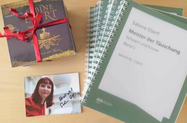 Auf einem Tisch liegt ein Hörbuch-Paket von Sabine Ebert mit roter Schleife, eine Autogramm-Karte der Autorin und ein Roman in Großdruck