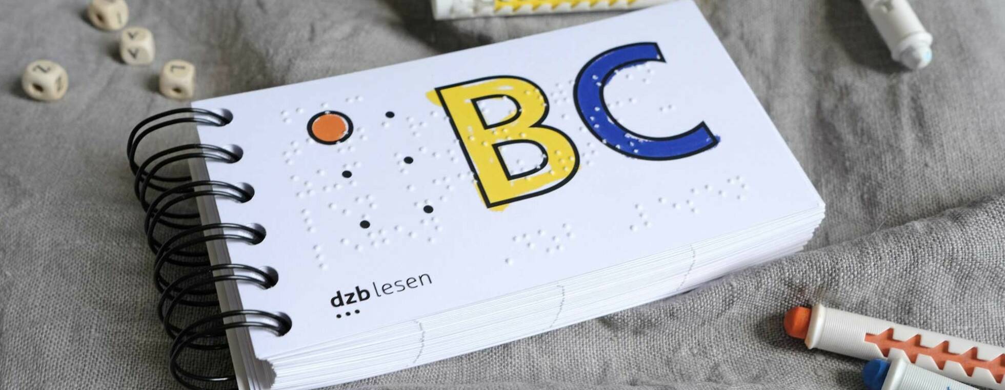 Ansicht des ABC-Buches mit Ringbindung. Auf dem Titel sind ein rotes A in Braille und ein gelbes B, ein lila C in Schwarzschrift mit Kontur. Neben dem Buch liegen Stifte und Buchstabenwürfel