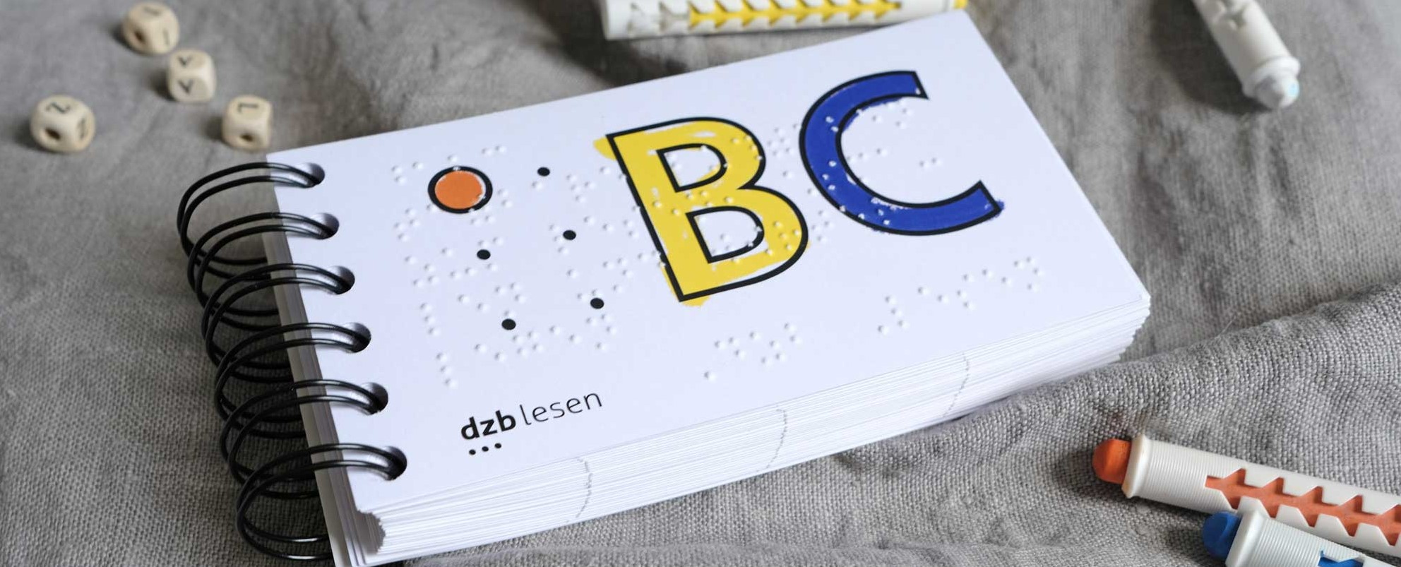Ansicht des ABC-Buches mit Ringbindung. Auf dem Titel sind ein rotes A in Braille und ein gelbes B, ein lila C in Schwarzschrift mit Kontur. Neben dem Buch liegen Stifte und Buchstabenwürfel