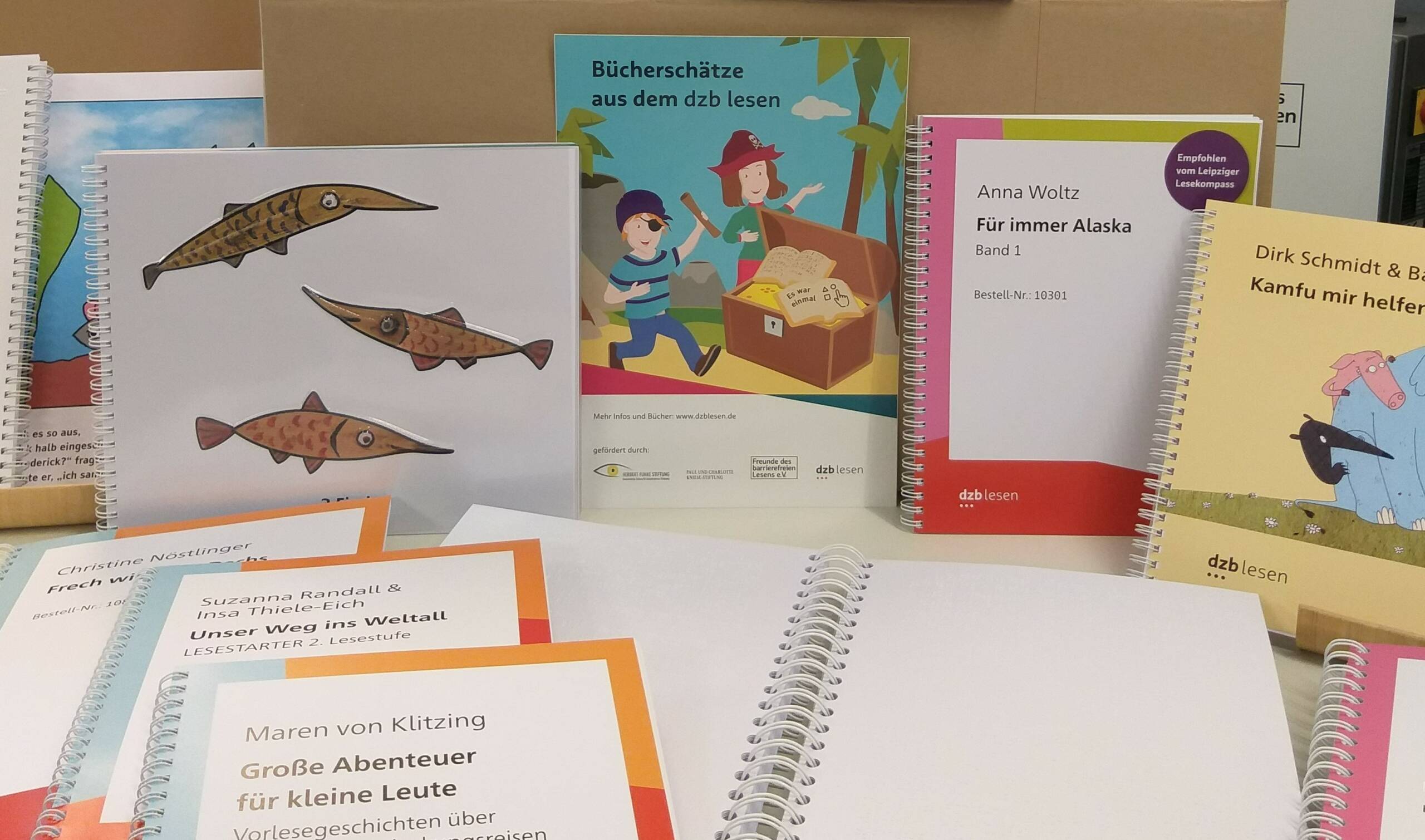 Foto: Viele bunte Kinderbücher liegen teilweise aufgeschlagen vor, an und auf einer Kiste. Im Hintergrund zwei Banner mit den Logos von FV und dzb lesen sowie dem Leseraben Oskar.