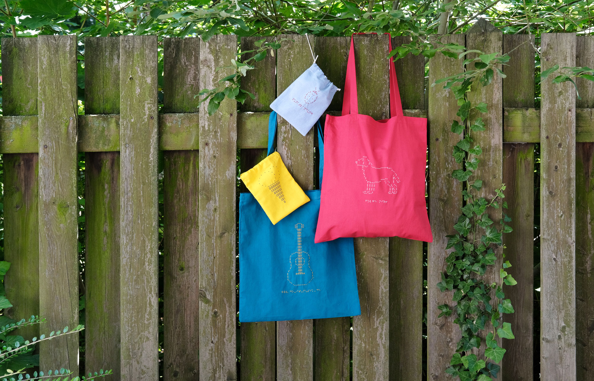 An einem Zaun hängen vier bunte Taschen und Beutel mit Braille-Stickerei