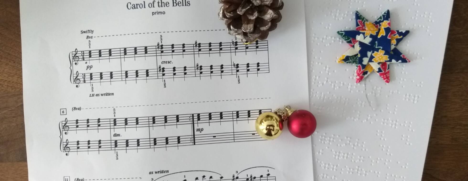 Zwei Notenblätter in Schwarzdruck und Braille von "Carol of the Bells" mit Tannenzapfen, Stern und Glöckchen