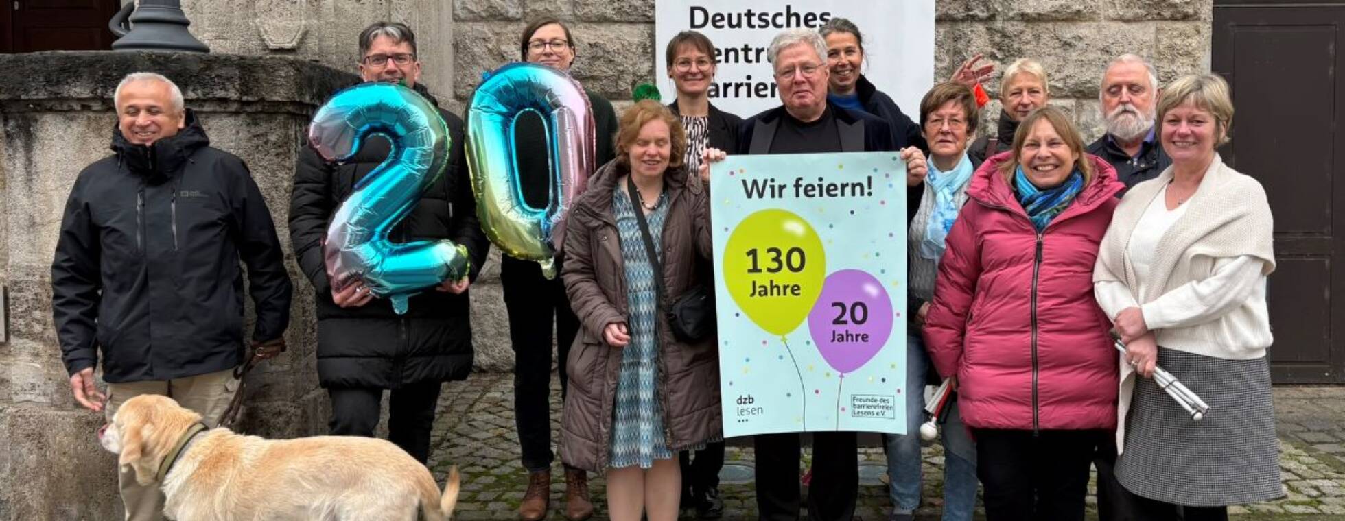 Foto einer Gruppe von 12 Menschen und einem Hind vor dem dzb lesen. In der Mitte wird das Jubiläumsplakat mit Luftballons gehalten, rechts im Bild zwei riesige Luftballons in Form einer 20.