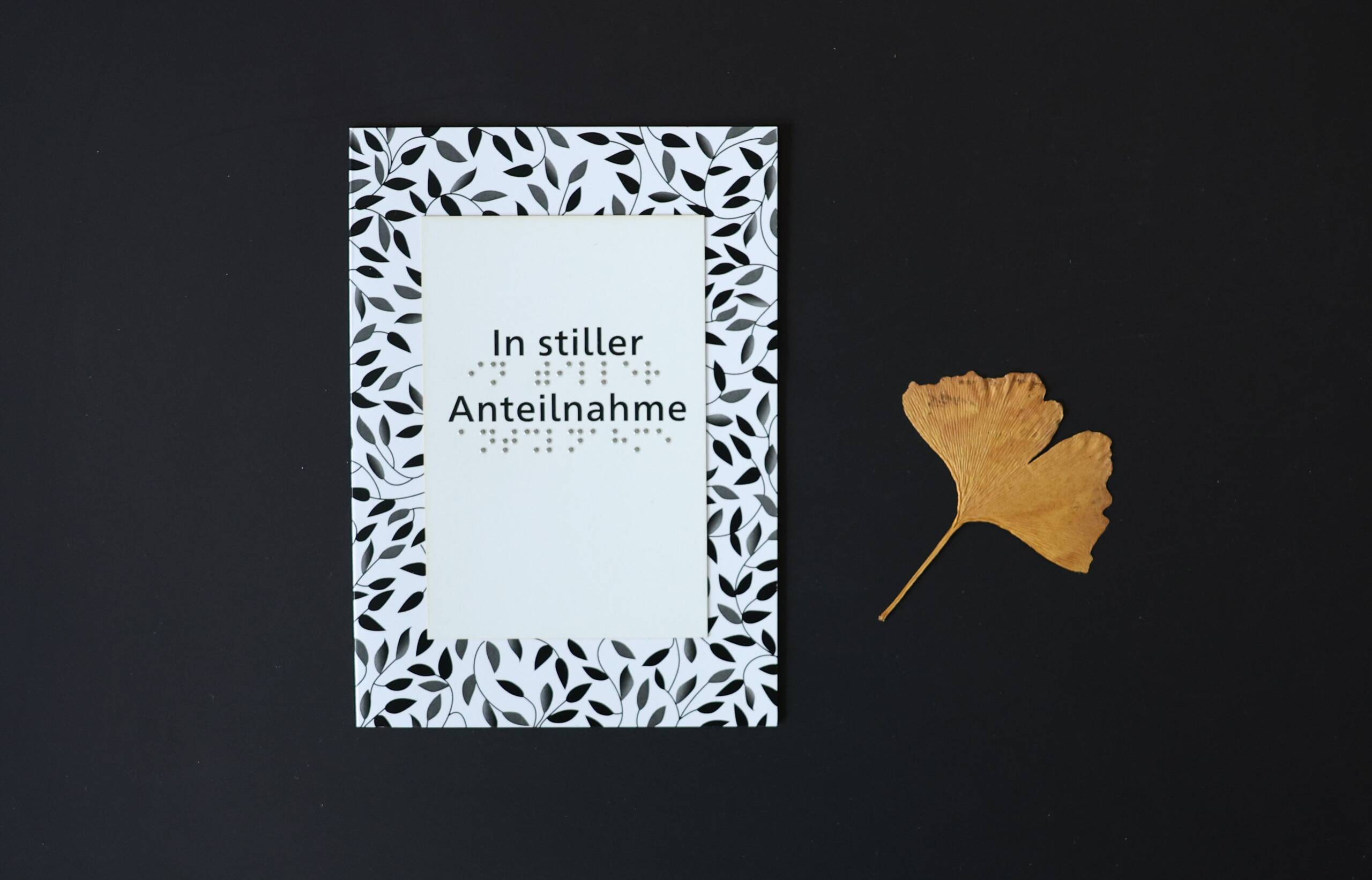 Eine schwarz-weiß gemusterte Trauerkarte in Schwarzdruck und Braille vor schwarzem Hintergrund mit einem goldenen Gingkoblatt rechts daneben. Auf der Karte steht der Text "In stiller Anteilnahme"