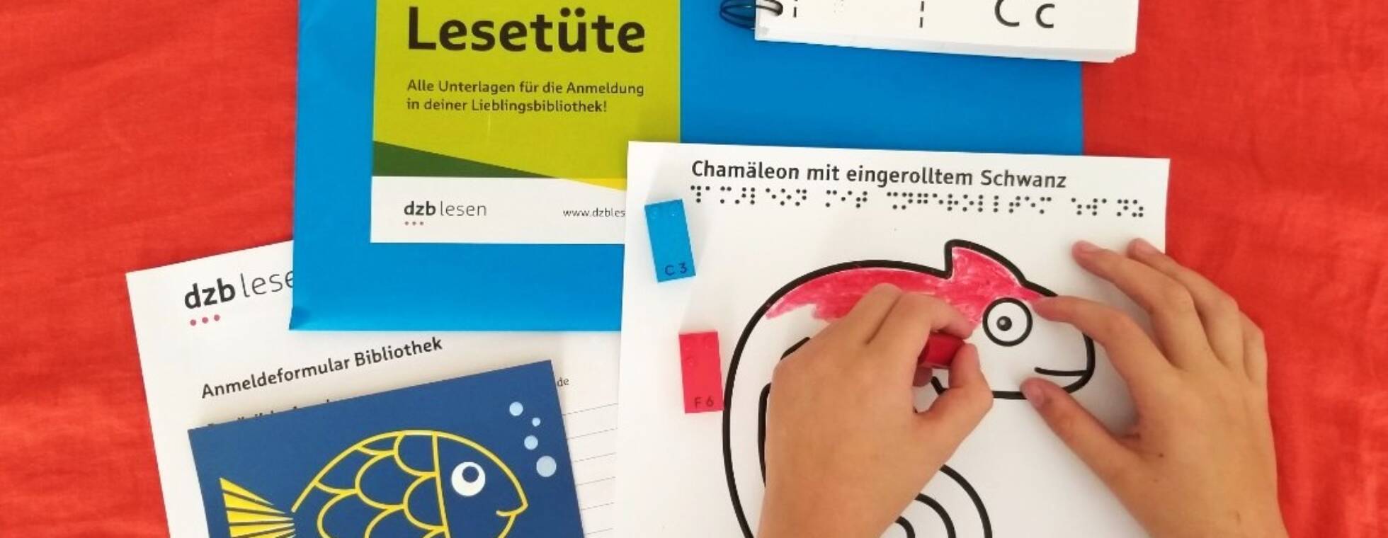 Der Inhalt der Lesetüte ausgebreitet auf rotem Grund: zwei Kinderhände malen ein Reliefbild eines Chamäleons aus.