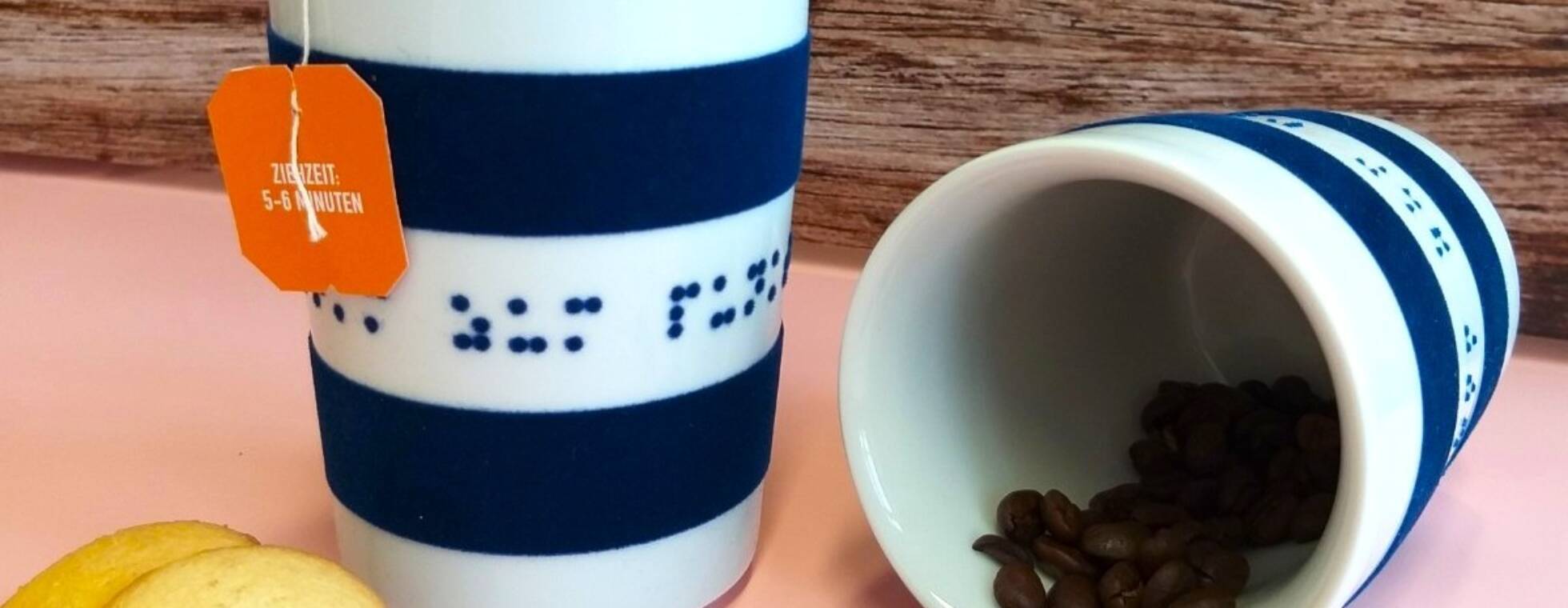 Zwei weiß-blaue Porzellanbecher, einer mit Kaffeebohnen, einer mit Teebeutel und Keksen vor einer Holzwand