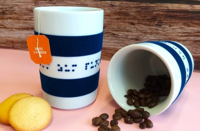 Zwei weiß-blaue Porzellanbecher, einer mit Kaffeebohnen, einer mit Teebeutel und Keksen vor einer Holzwand