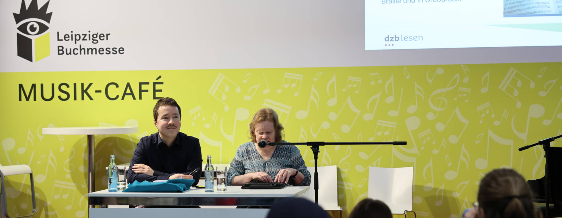 Anja Lehmann am Mikrofon und René Holzhauer vor der gelben Kulisse des Musik-Cafés der Buchmesse. Im Vordergrund das Publikum.