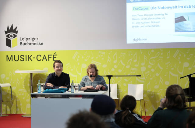 Anja Lehmann am Mikrofon und René Holzhauer vor der gelben Kulisse des Musik-Cafés der Buchmesse. Im Vordergrund das Publikum.