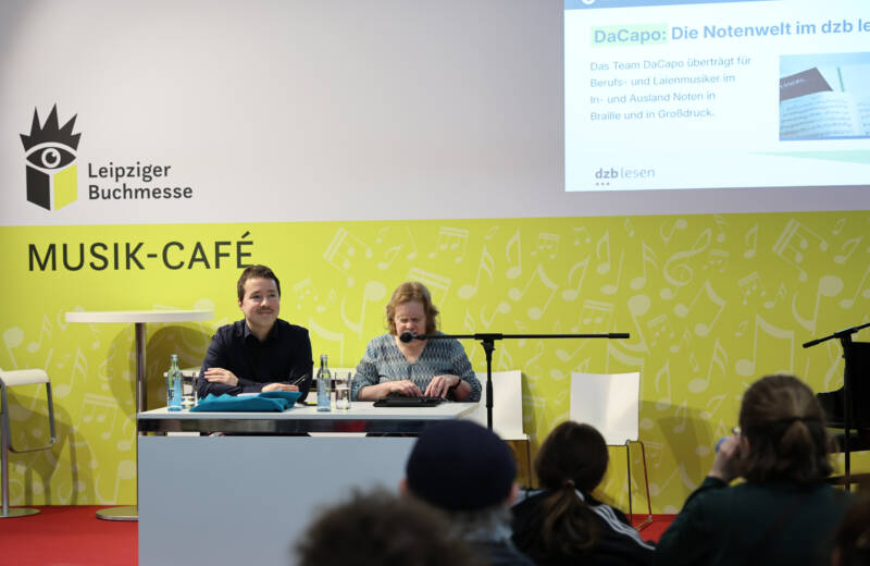 Anja Lehmann am Mikrofon und René Holzhauer vor der gelben Kulisse des Musik-Cafés der Buchmesse. Im Vordergrund das Publikum.