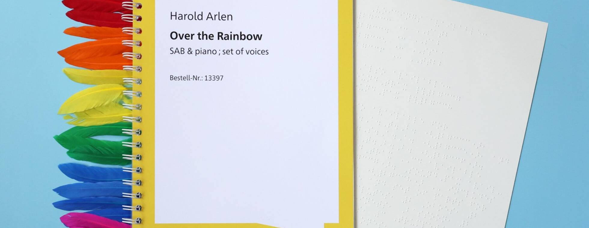 Ein Ringbuch, aus dem eine Braillenotenseite ragt vor blauem Hintergrund. Links vom Buch liegen Federn in den Farben des Regenbogens. Text auf dem Titel: Text "Harold Arlen, Over the Rainbow. SAB piano. set of voices"