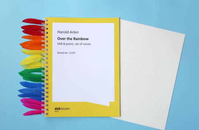 Ein Ringbuch, aus dem eine Braillenotenseite ragt vor blauem Hintergrund. Links vom Buch liegen Federn in den Farben des Regenbogens. Text auf dem Titel: Text "Harold Arlen, Over the Rainbow. SAB piano. set of voices"